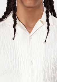 Biała koszula zapinana na guziki z fakturowaną, pomarszczoną powierzchnią. Posiada klasyczny kołnierzyk i małe zapięcie na guzik, co podkreśla jej elegancki wygląd.