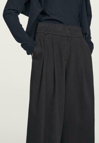 Pantaloni a vita alta, plissettati, in tessuto grigio scuro con una texture liscia. Presentano tasche laterali e una vestibilità sagomata.