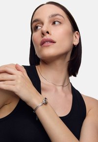 Srebrna bransoletka z motywem węzła. Gładka metalowa tekstura, smukły kształt, noszona na nadgarstku. Tło w jasnoszarym kolorze. Prosta, minimalistyczna choker.