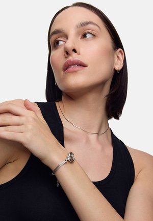 Srebrna bransoletka z motywem węzła. Gładka metalowa tekstura, smukły kształt, noszona na nadgarstku. Tło w jasnoszarym kolorze. Prosta, minimalistyczna choker.