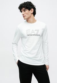 Valge pikkade varrukatega särk musta "EA7 EMPORIO ARMANI" logoga. Sile tekstuur, keha jälgiv lõige, sobitatud mustade pükstega.