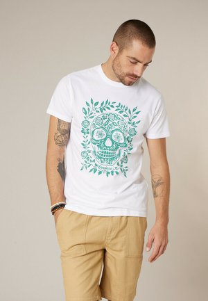 Homme aux cheveux courts et tatouages, portant un t-shirt blanc avec un motif de crâne floral vert et un short beige, regardant vers le bas les mains dans les poches.