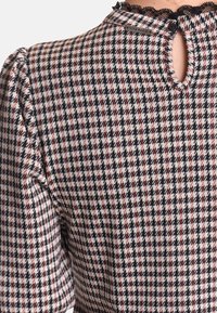 Svartvit hundtandsmönstrad topp med subtila röda accenter. Puffärmar och spetskant vid kragen, med en nyckelhålsöppning bak.