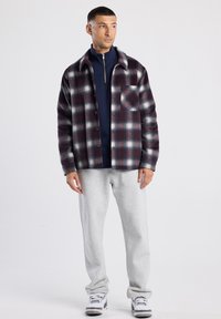 Ruudullinen flanellipaita tummanpunaisena, valkoisena ja laivastonsinisenä, kauluksella ja taskulla, puettuna laivastonsinisen vetoketjullisen paidan ja harmaiden verkkareiden päälle.