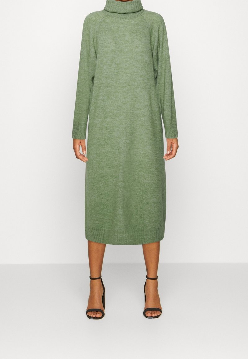 Femme portant une robe pull à col roulé verte, longue jusqu'aux genoux, à manches longues, avec des escarpins noirs à bride à la cheville, debout devant un fond uni.