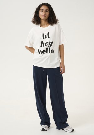 T-shirt blanche avec un texte noir en gras "hi hey hello", associé à un pantalon ample bleu marine et des baskets grises. Tenue décontractée et tissu doux.