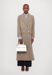 Manteau long beige à double boutonnage avec des boutons métalliques, porté sur une chemise rayée bleu et blanc. Tenant un petit sac à main blanc. Chaussures marron.
