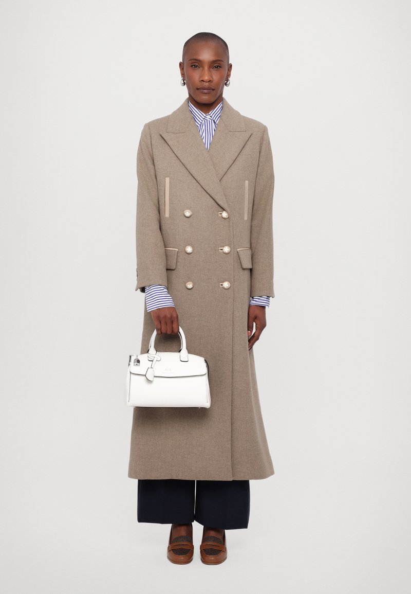 Manteau long beige à double boutonnage avec des boutons métalliques, porté sur une chemise rayée bleu et blanc. Tenant un petit sac à main blanc. Chaussures marron.