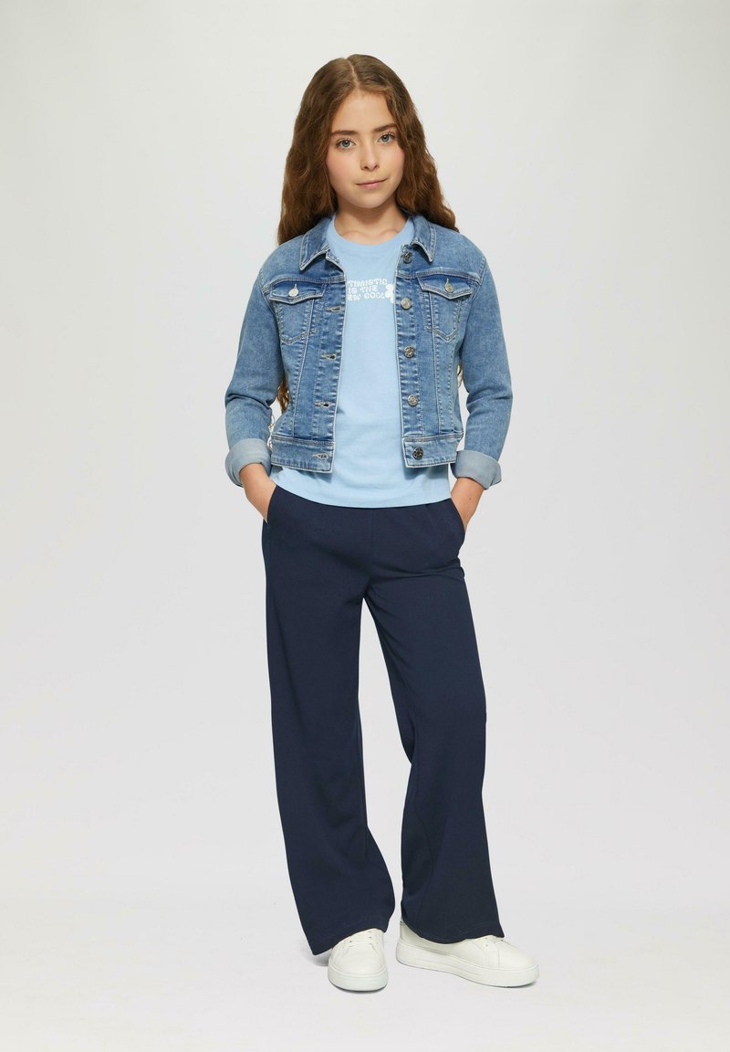 Jeansjack met een lichtblauw t-shirt en donkerblauwe wijde broek. Witte sneakers maken de outfit compleet. Lang golvend haar omkadert het gezicht.