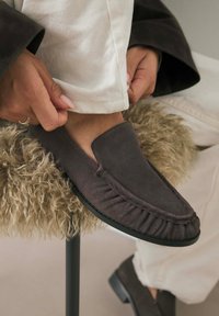 Hallhalli siidist lipatsid, millel on frantsi aktsendid ja must kummitald, kantud jalal, puhkamas tekstuurilisel beežil ja kreemikas pinnal.