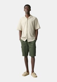 Camicia beige a maniche corte con bottoni e motivo testurizzato, abbinata a pantaloni corti verde oliva e scarpe slip-on beige.