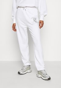 Persoon in witte sweatpants met een zilveren logo op de dij, gecombineerd met zilveren en witte sportieve sneakers, staande tegen een effen achtergrond.