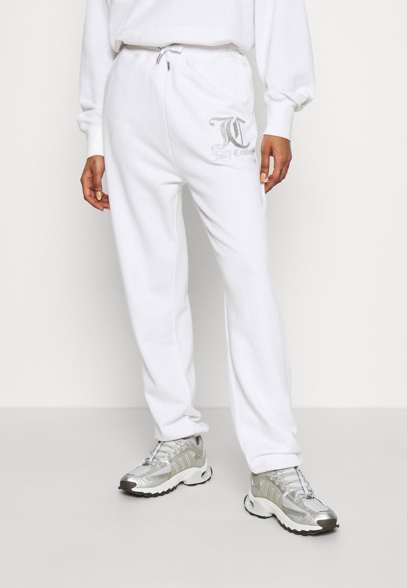 Persoon in witte sweatpants met een zilveren logo op de dij, gecombineerd met zilveren en witte sportieve sneakers, staande tegen een effen achtergrond.