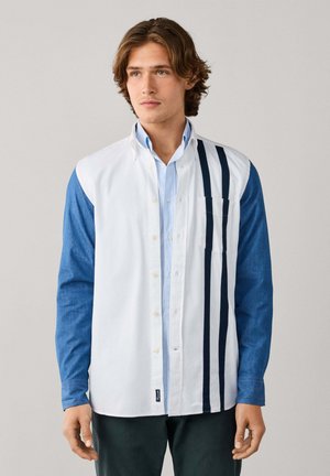 Hombre de pie, vistiendo una camisa abotonada con parte delantera blanca, mangas azules y tres rayas verticales azul marino en el lado derecho. Fondo neutro.