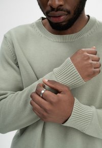 Hellgrüner Strickpullover mit gerippten Bündchen, getragen von einer Person, die ihre Arme hält. An einer Hand befindet sich ein silberner Ring. Glatte Textur, lässiges Design.