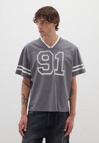 Camiseta de jersey gris con cuello en V y rayas blancas en las mangas. Presenta el número "91" desgastado impreso en la parte frontal en blanco.