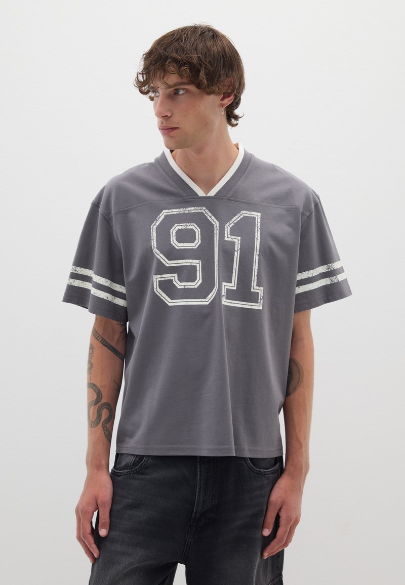Camiseta de jersey gris con cuello en V y rayas blancas en las mangas. Presenta el número "91" desgastado impreso en la parte frontal en blanco.