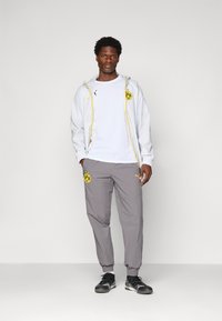 Homme debout portant un pantalon de sport gris, un t-shirt de sport blanc, une veste grise claire avec des détails jaunes et des baskets noires sur fond uni.