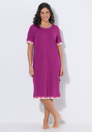 Frau lächelt, steht barfuß und trägt ein knielanges violettes Kleid mit rosa Spitzenbesatz an Ärmeln und Saum vor einem eintönigen weißen Hintergrund.