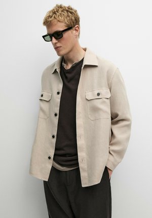 Lyshåret mand iført sorte solbriller, beige skjortejakke med knapper over en sort T-shirt og sorte bukser, stående mod en ensfarvet hvid baggrund.