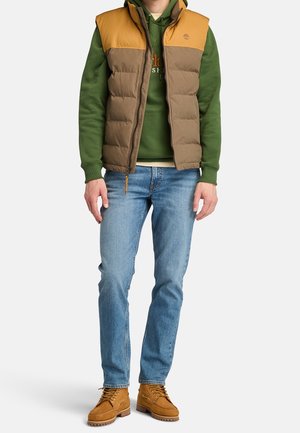 Gilet imbottito marrone e arancione con chiusura a zip, abbinato a un hoodie verde, jeans blu e stivali marroni con lacci.