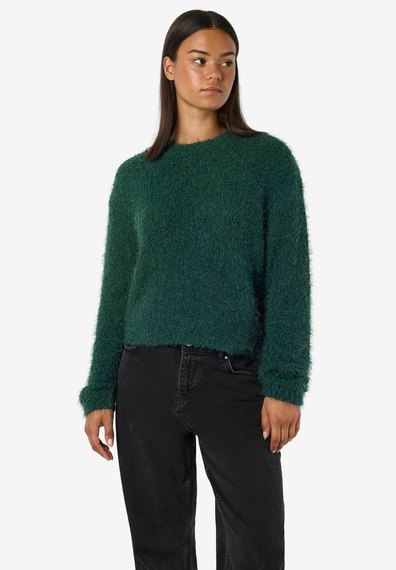 Zelen pulover z mehkimi dlačicami in klasičnim krojem, dolgimi rokavi in ribastimi manšetami, v kombinaciji s črnimi džinsom, ki ima mehak občutek.