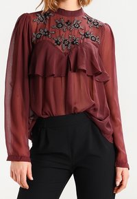 Blouse transparente couleur bordeaux avec volants, encolure ornée de perles florales, manches longues et taille froncée, associée à un pantalon noir.