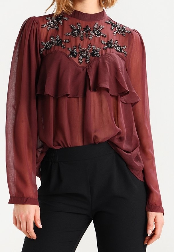Blouse transparente couleur bordeaux avec volants, encolure ornée de perles florales, manches longues et taille froncée, associée à un pantalon noir.