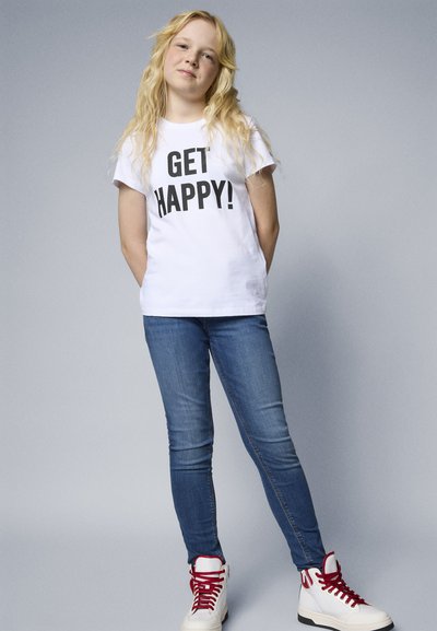 Vit bomull T-shirt med fet svart text "BLI GLAD!" tillsammans med blå jeans och vita högtoppsneakers med röda snören.