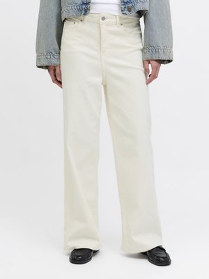 Person iført løstsiddende hvide jeans, sorte sko, hvid skjorte og cropped blå denimjakke mod ensfarvet baggrund.