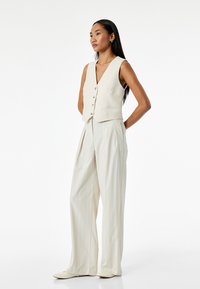 Top senza maniche color crema con scollatura a V profonda e chiusura a bottoni, abbinato a pantaloni larghi a righe sottili grigi chiari e scarpe a punta abbinate.