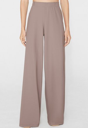Mujer con pantalones taupe de cintura alta y pierna ancha con cremallera lateral, de pie frente a un fondo claro y liso.