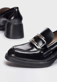 Mocasines de cuero negro con un acabado brillante, featuring un amplio detalle de hebilla plateada y un tacón negro grueso y texturizado.