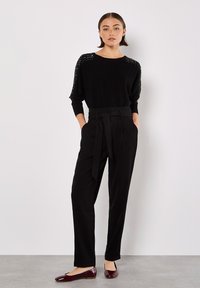 Schwarzes Strickoberteil mit Nieten an den Schultern, kombiniert mit hoch taillierten schwarzen Hosen mit gebundenem Gürtel und glänzenden burgunderfarbenen Ballerinas.