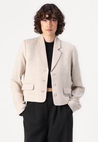 Beige színű, texturált anyagból készült rövid blézer, kivágott hajtókával, három gombbal és két elöl található zsebbel; fekete felsőre húzva.