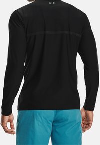 Homme portant un t-shirt d'athlétisme noir ajusté à manches longues avec un logo dans le haut du dos et un short texturé bleu vif, se tenant avec les bras détendus.
