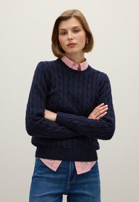 Marineblauer Kabelstrickpullover mit geripptem Saum und Bündchen, über einem rot-weiß gestreiften Hemd mit Kragen getragen, kombiniert mit blauen Jeans.