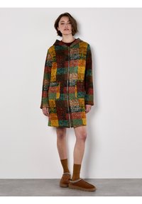 Kurtka z kapturem, zapinana na zamek, z kolorowym wzorem w kratę w ziemistych odcieniach zieleni, czerwieni, żółci i brązu. Dwie widoczne przednie kieszenie.