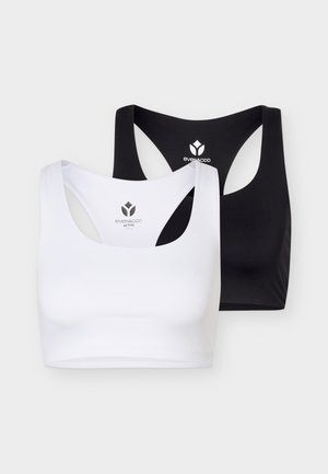 Fit persoon die frontaal staat en een zwarte sportbeha, bordeauxkleurige leggings en zwarte sportschoenen draagt op een lichte achtergrond.