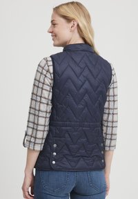 Gilet trapuntato blu navy con motivo a zigzag, dotato di colletto alto, bottoni laterali e vita cinch. Indossato sopra una camicia a quadrati.