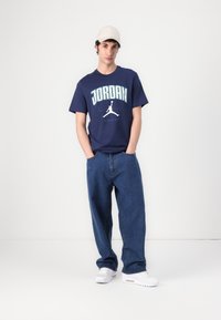 Marineblå T-shirt med "JORDAN" grafik og silhuetlogo, parret med løse blå jeans og hvide sneakers. En beige caps fuldender looket.