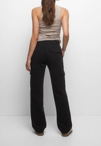 Žena s dlouhými hnědými vlasy má na sobě béžový top bez rukávů a černé široké kalhoty s cargo kapsami, stojící na jednobarevném bílém pozadí.