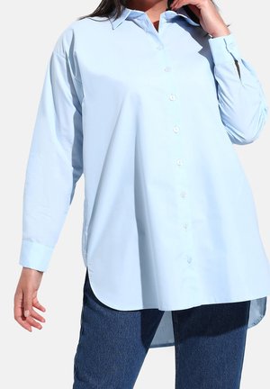 Mujer con una camisa holgada de botones de color azul claro y mangas largas, combinada con vaqueros azul oscuro, posando con una mano cerca de su cara.