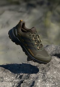 Olivengroene trailrunning schoenen gemaakt van synthetisch materiaal, met gaasaccenten, een gestructureerde zool en tan details, waaronder het logo en de veters.