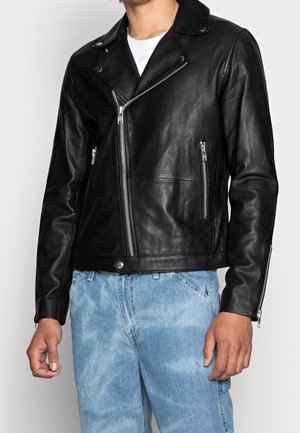 Mand iført sort læder bikerjakke med sølvfarvede lynlåse og lyseblå denimjeans, stående mod en ensfarvet baggrund.