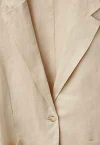 Beige linnen blazer met een geknipte revers, sluiting met één knoop en een subtiele textuur. Inclusief twee voorzakken. Eenvoudig en elegant ontwerp.