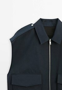 Granatowa bezrękawnik z wyraźnym kołnierzem, srebrnym zamkiem błyskawicznym i dwiema przednimi kieszeniami. Posiada kontrastowe obszycie na ramionach oraz detal z guzikami.