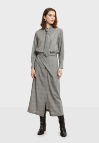 Siva karirana haljina s detaljem vezanja oko vrata, dugim rukavima i prorezom sprijeda, uparena s crnim čizmama do koljena. Glatka tekstura tkanine.