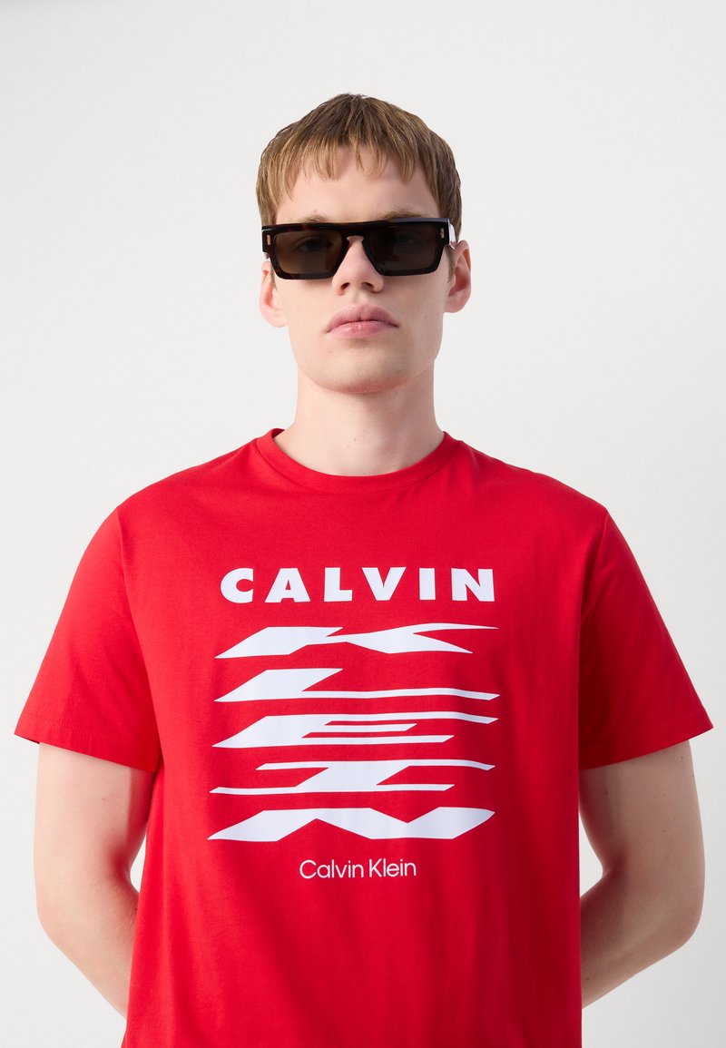 T-shirt en coton rouge arborant un texte blanc en gras dans une police moderne et des motifs abstraits. Des lunettes de soleil rectangulaires noires complètent le look.