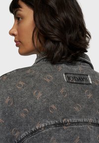 Giacca di jeans grigia con un motivo ripetuto di logo marrone chiaro, caratterizzata da una finitura testurizzata e un'etichetta vicino al colletto.